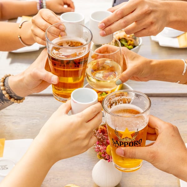 Multiple hands clinking glasses of beer, wine, and sake cups in a celebratory toast at Kombu Sushi.