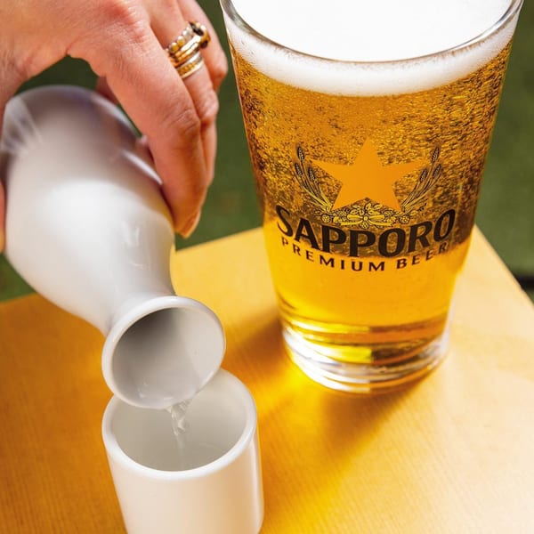 A hand pours sake from a white carafe into a small cup next to a glass of Sapporo beer.