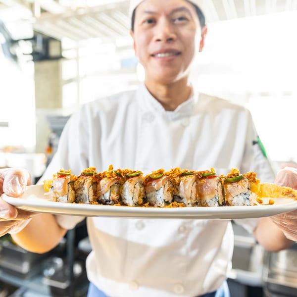 Kombu Sushi chef presenting a long plate of sushi rolls topped with crispy flakes and jalapeños.