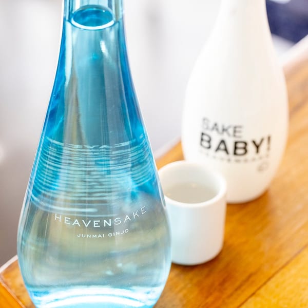 A blue bottle of "Heaven Sake" and a white bottle of "Sake Baby" with a small white cup.